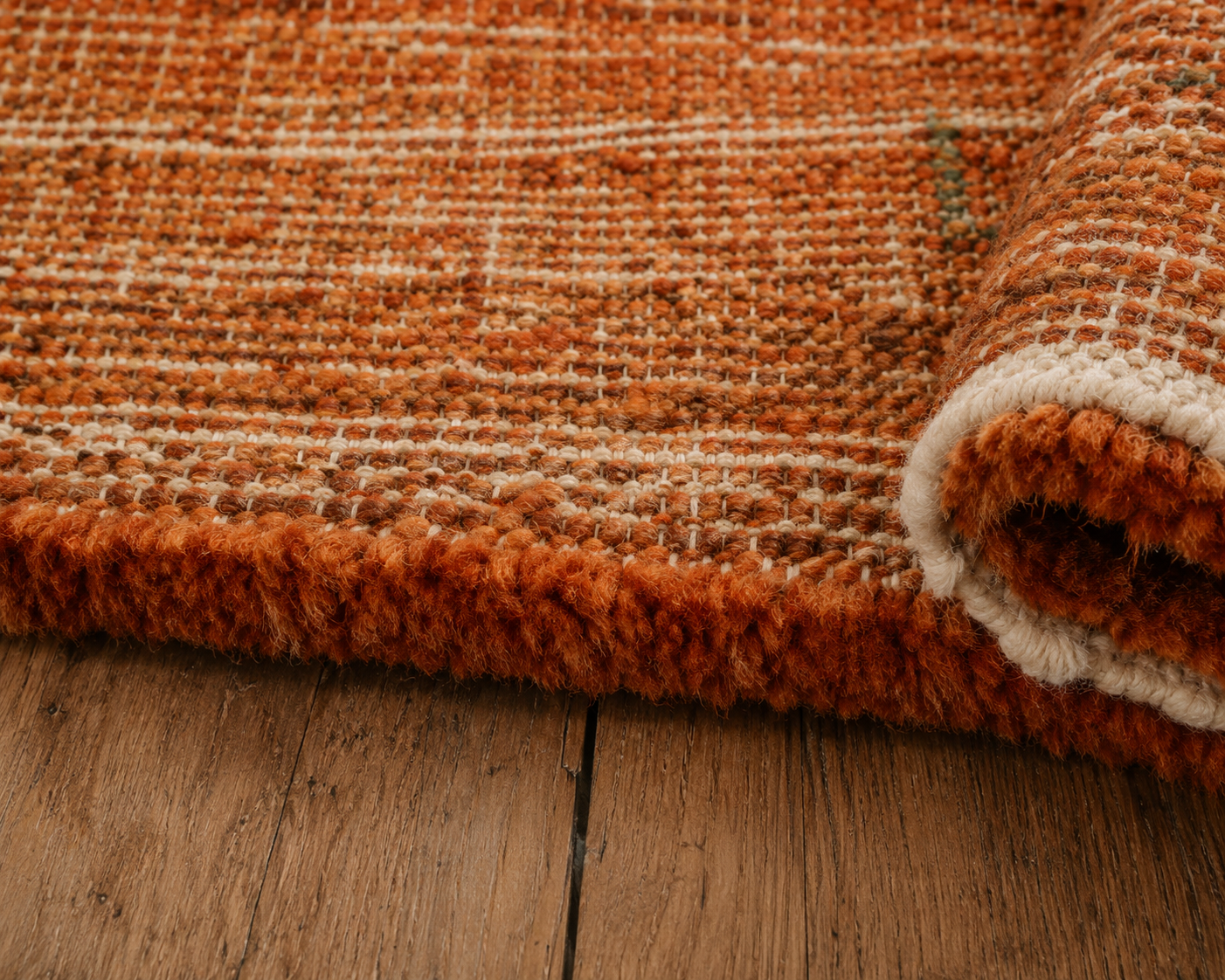A close-up of a textured woven rug in rust orange and cream, partially rolled up on a wooden floor.