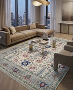 A large traditional-style area rug with an intricate floral and medallion pattern in cream, blue, red, and beige tones, styled in a modern living room setting with a sectional sofa, glass coffee table, and city view windows.